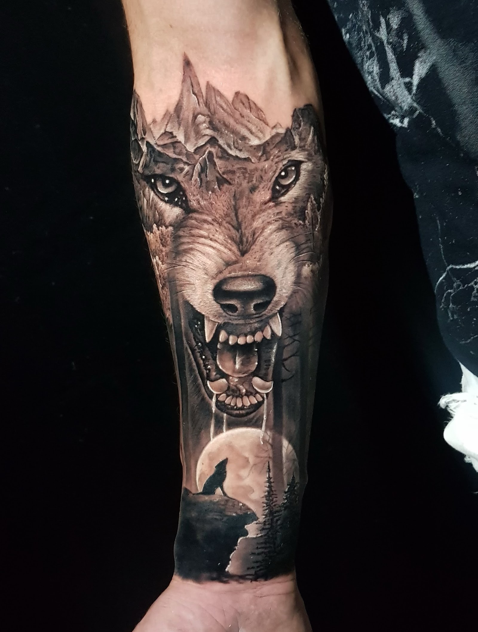 Tatouage r&eacute;aliste noir et gris d'un loup avec montagnes et pleine lune sur un avant-bras